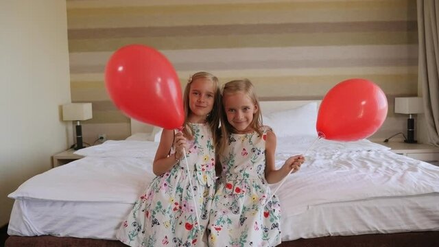 Two twin sisters are sitting on a bed in a bedroom with red balloons. Portrait of twin girls in a hotel with identical balloons.