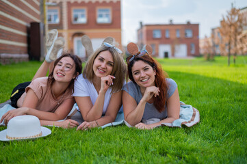 Three beautiful Caucasian young women lie on the lawn in the countryside. The blonde brunette and the redhead are resting in the park on the green grass. The friends had an outdoor picnic.