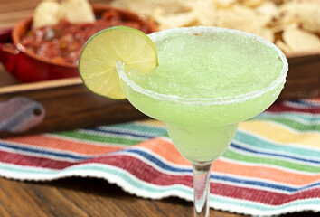 Frozen margarita garnished with lime slice, out of focus red salsa and corn chips in background