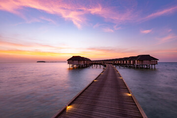 Obraz premium Sunset on Maldives island, luxury water villas resort and wooden pier. Beautiful sunset sky and clouds and tropical beach background for summer vacation holiday and travel concept banner 