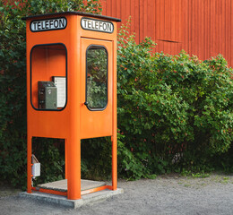 Fototapeta premium Old swedish orange telephone box in front of wooden building and hedges