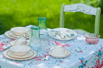 Outdoor vintage table setting with floral tablecloth, fine china and silverware arranged in a shabby chic manner including antique distressed chair, 