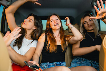 Three different beautiful female friends sit at vehicle back seat, listen music songs, singing and...