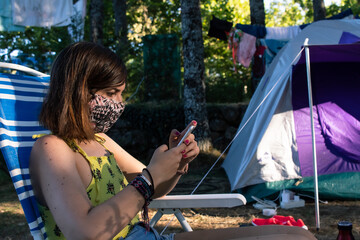 Attractive young female looking at her smartphone while sitting on a chair in camping and wearing a facemask