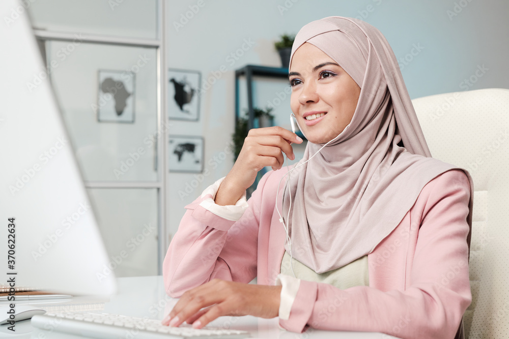 Aufkleber Smiling young Muslim manager in pink hijab using ear pieces while communicating via video conferencing app on computer in office