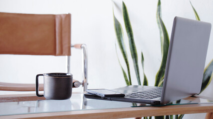 Trabajar desde el hogar. Plano frontal en un interior iluminado y blanco de una taza negra de cafe junto a una notebook y un teléfono celular en un pequeño escritorio. 