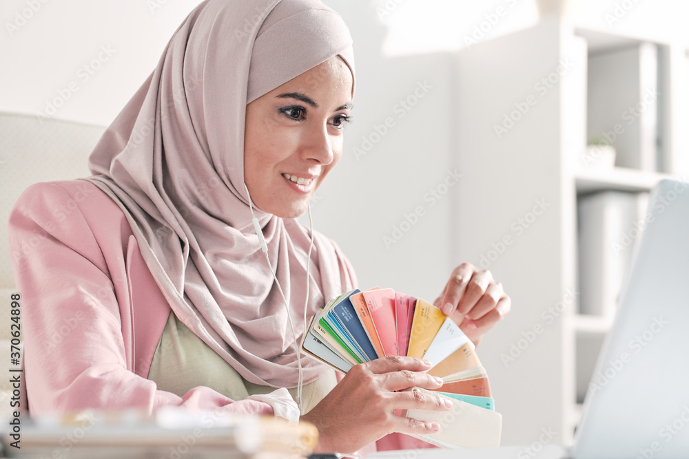 Aufkleber Smiling Muslim designer in pink hijab showing color swatch to customer while talking to him via video conferencing app