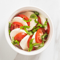 Fatlay of Red tomatoes, green basil and white buffalo mozzarella for a Caprese Salad on a white plate/bowl on a marble table and background