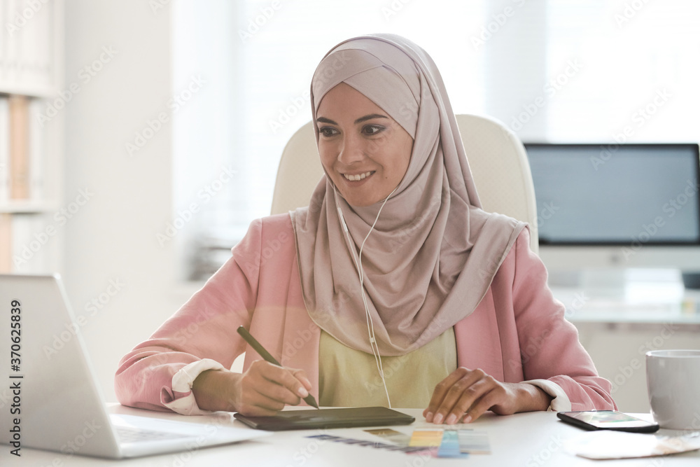 Poster Smiling young Muslim graphic designer in hijab sitting at desk and using drawing pad while working on web project
