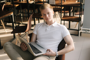 Serious young businessman wearing fashion casual clothing sitting on chair and holding laptop in his hand, looking at camera. Successful business man is working on laptop computer in start-up office.