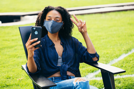 Black Woman Sitting In Patio Chair  With A Face Mask Taking A Selfie During Covid 19