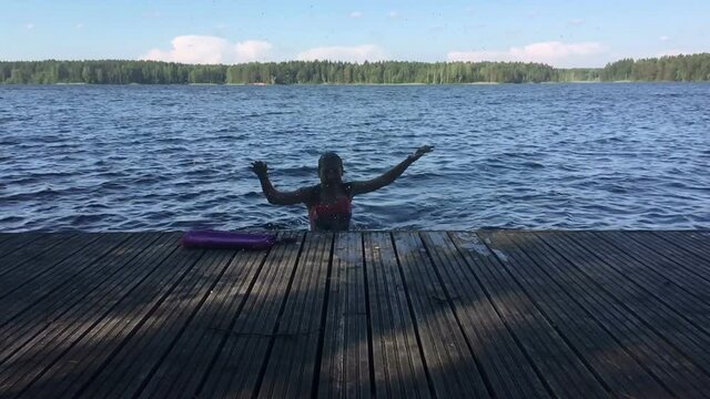 Preteen girl jumping from water and plashing, slow motion view. Wooden pier is on forest lake