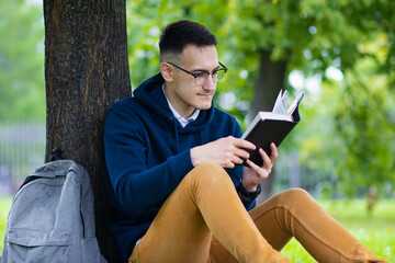 Obraz premium Young concentrated guy in glasses, student reading an interesting book, leaning at a tree, relaxing in summer park with backpack. Handsome smart intelligent man studying outdoors.