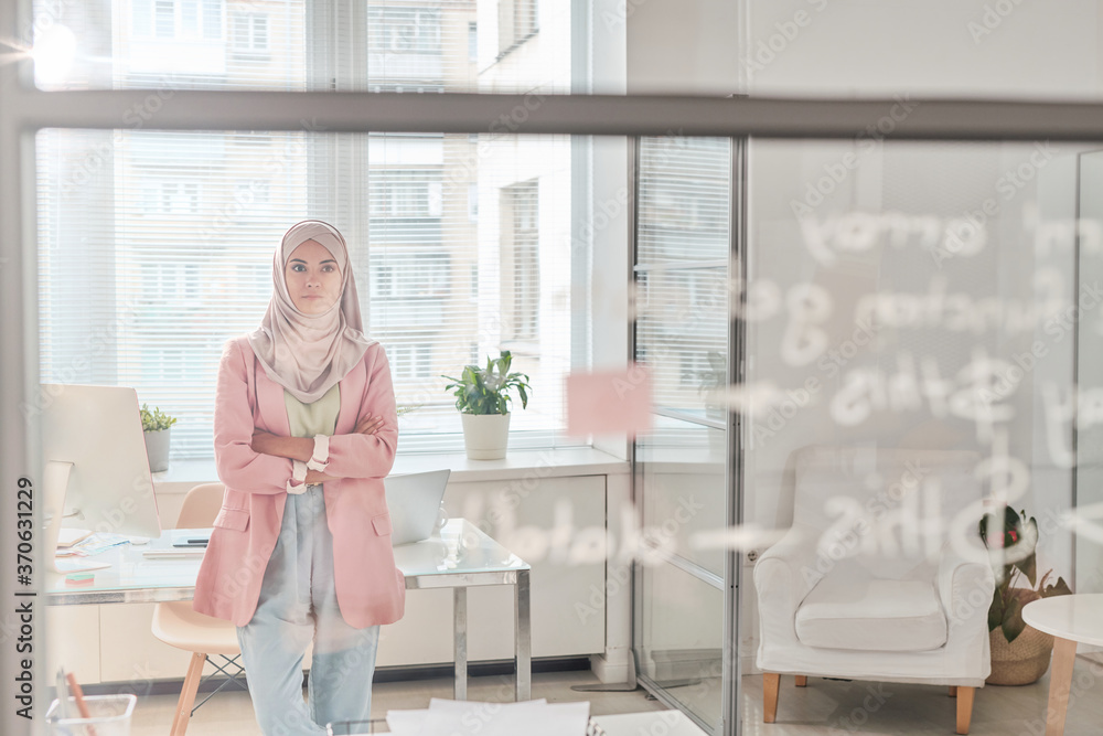 Poster Serious young Muslim coder in hijab standing with crossed arms in office and looking at board while thinking about new app