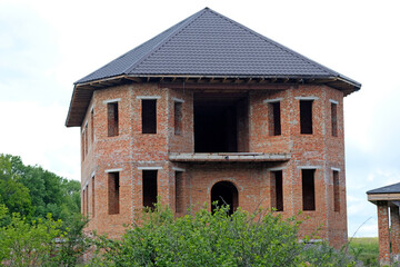 Fototapeta na wymiar Facade of an unfinished two-story house with no red brick windows. Large house with rounded corners and a large terrace. The process of building a house on a large green plot near the forest.