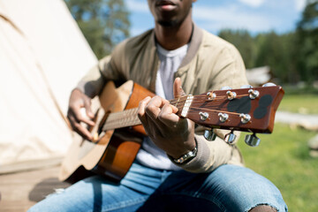 Close-up of young Black man sitting on wooden porch of camping tent and playing guitar outdoors