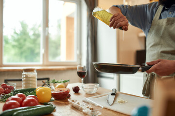 Cropped shot of man, cook in apron pouring oil on the pan for preparing dinner, standing in the...