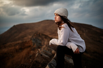 Young beautiful woman in a white sweater stands on top of a mountain and enjoys the scenery. Young woman standing on cliff's edge and looking to a sky - 370635616