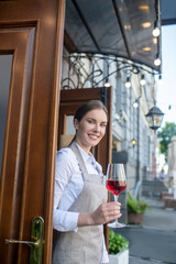 Smiling cute waitress in grey apron holding glass of wine