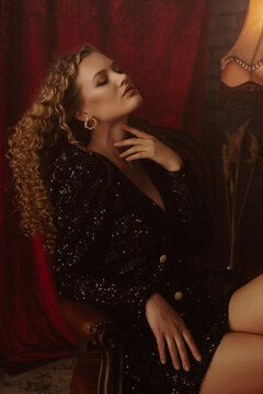 Luxury Blonde Woman With Long Curly Hair, Wearing Trendy Sequin Mini Dress With Big Shoulders Posing In Dark Retro, Vintage Interior. Model Sitting On The Chair
