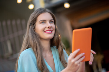 Romantic female in blue dress sitting outside, holding orange tablet, smiling