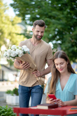 Young female sitting at table, checking her phone, bearded male coming to her with flowers