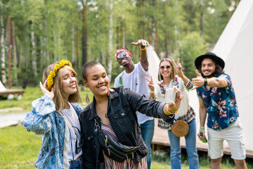 Fototapeta premium Positive black woman with long earrings photographing with friend in wreath on smartphone while their friends making cool gestures in background