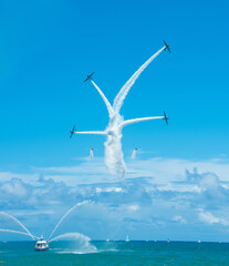 Airplanes performing a starburst maneuver above the ocean, paralleling a ship spouting water in...