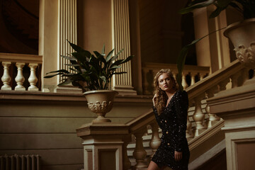 Luxury blonde woman wearing trendy sequin mini dress walking up stairs in beautiful old building. Film grain effect. Copy, empty space for text