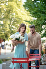Naklejka premium Bearded male pulling red chair for female with bouquet of white flowers, helping her to sit