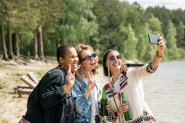 Cheerful attractive young multi-ethnic women with beer bottles taking selfie together on beach