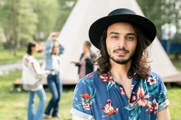 Portrait of content handsome Jewish guy in black hat and shirt with flower print resting with friends at forest campsite