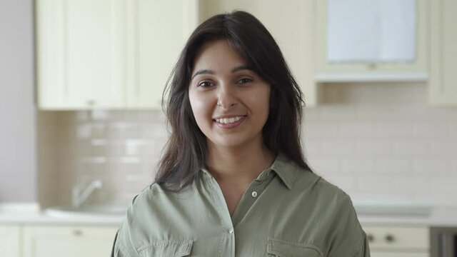 Smiling young pretty indian ethnicity woman looking at camera alone at home in kitchen. Happy beautiful millennial hindu lady housewife in India indoors, close up face front headshot portrait.
