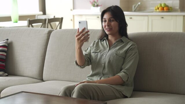 Young happy indian woman sitting on sofa at home holding phone video calling distance friend dating online in mobile app using smartphone videochat application talking communicating in video chat.