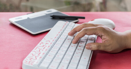 Woman's hand typing on a white keyboard with red letters, with graphic tablet in the background. On red table.