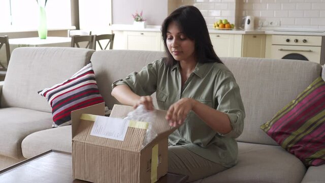 Happy indian young woman buyer opening parcel box at home. Smiling female customer shopper receiving online shop purchase buying clothes unpacking delivery postal shipping package gift sit on couch.