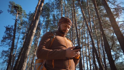 theme tourism and technology. Young caucasian man with beard and backpack. Hiking tourist in pine forest uses technology, hand holding mobile phone to touch the screen. Gps application orientation