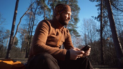 hiking nature travel. Caucasian male tourist with beard and backpack resting, break turned felled tree, stump in pine forest autumn in sunny weather. Uses technology hand smartphone touch screen