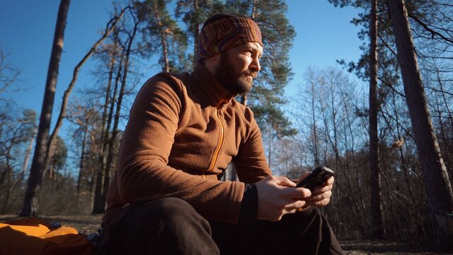 hiking nature travel. Caucasian male tourist with beard and backpack resting, break turned felled tree, stump in pine forest autumn in sunny weather. Uses technology hand smartphone touch screen