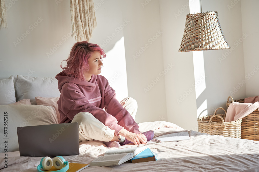 Wall mural Dreamy thoughtful teen hipster girl with pink hair wears hoodie sits in bed with laptop studying thinking of ideas, good future plans, looking away, reflecting, dreaming, lost in thoughts in bedroom.