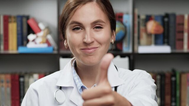 Doctor thumb up. Friendly woman doctor in white coat and stethoscope in office looks at camera and raises her hand, shows thumb up, nods and smiles approvingly