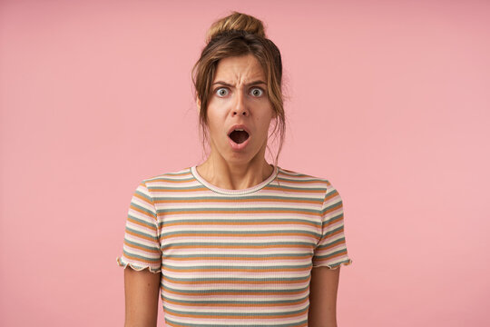 Confused young attractive brown haired woman keeping her mouth wide opened while looking indignantly at camera, posing over pink background in striped t-shirt