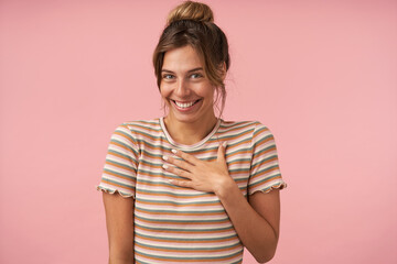 Portrait of young beautiful brown haired woman with natural makeup looking cheerfully at camera with wide smile while posing over pink background in casual wear