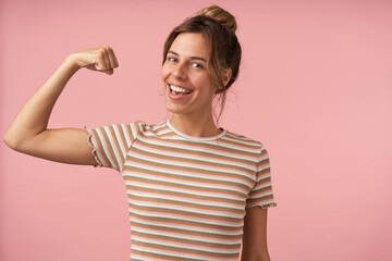 Studio photo of charming young brunette female dressed in beige striped t-shirt raising hand while demonstrating her power and smiling gladly at camera, isolated over pink background
