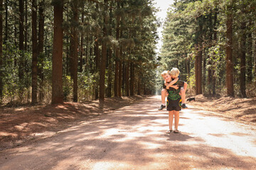 Fototapeta premium Boy giving his little brother a piggy back on dirt road among rows of tall pine trees