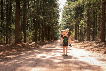 Boy giving his little brother a piggy back on dirt road among rows of tall pine trees