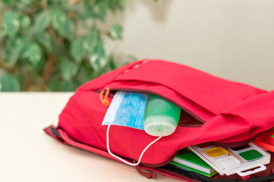 Back to school. Education concept. Red satchel with protective medical mask, antibacterial hand cleaning gel (hand rub), school supplies on light background. Folding school supplies into a backpack.