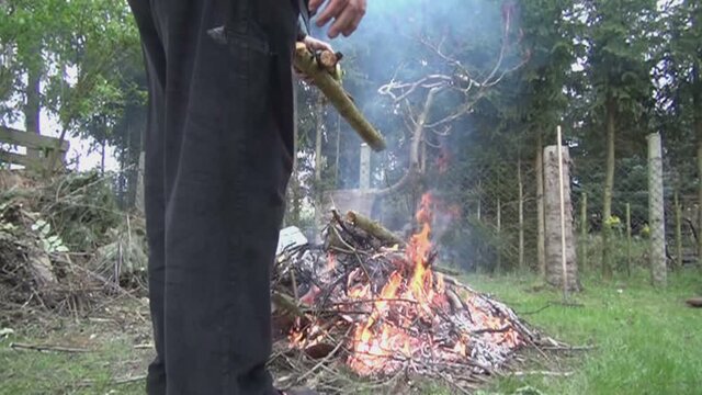 Gro&szlig;er robuster Mann wirft einen Ast auf ein lichter loh brennendes Osterfeuer. Zeitlupenaufnahme eines Stocks, der auf das Lagerfeuer geschmissen wird. Glut spritzt, Rauch entsteht, Holz kracht. Fire