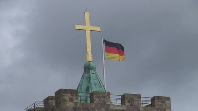 Die deutsche Flagge: "Schwarz, rot, gold" weht in Zeitlupe auf einer Turmspitze neben einem goldenen Kreuz. Dunkle Wolken im Hintergrund machen eine d&uuml;stere Stimmung. Die Wartburg in Eisenach.