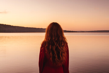 Young ginger woman in red dress at sunset. Back view. River or lake on background, pink sky.